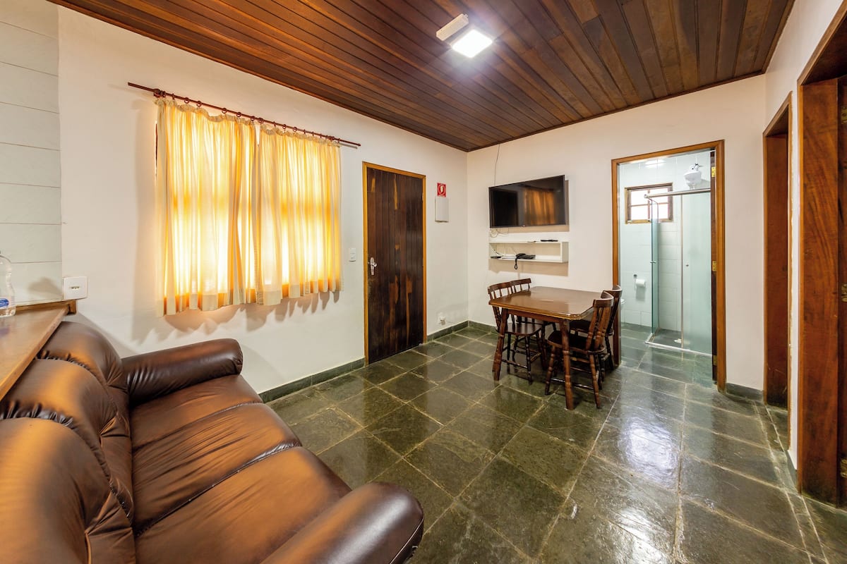 A living room with a brown leather sofa, a dining table with chairs, a TV mounted on the wall, and a door leading to a bathroom.