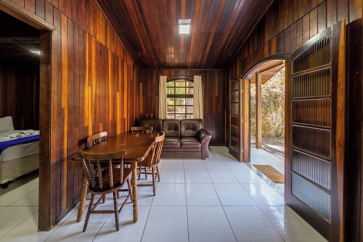 A wooden interior with a dining table and chairs, a sofa, and a window with curtains.