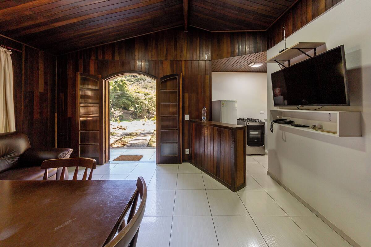 A spacious room with a wooden ceiling and walls, a dining area with a table and chairs, a kitchen area with a refrigerator and stove, and a large TV mounted on the wall.