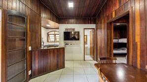 A wooden interior with a dining table and chairs, a kitchen area with a TV, and a bed in the background. - Hotel Fazenda da APM by Bhomy (Caieiras)