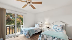 A bedroom with two beds, a ceiling fan, a window with a view of trees, and a wooden deck outside.