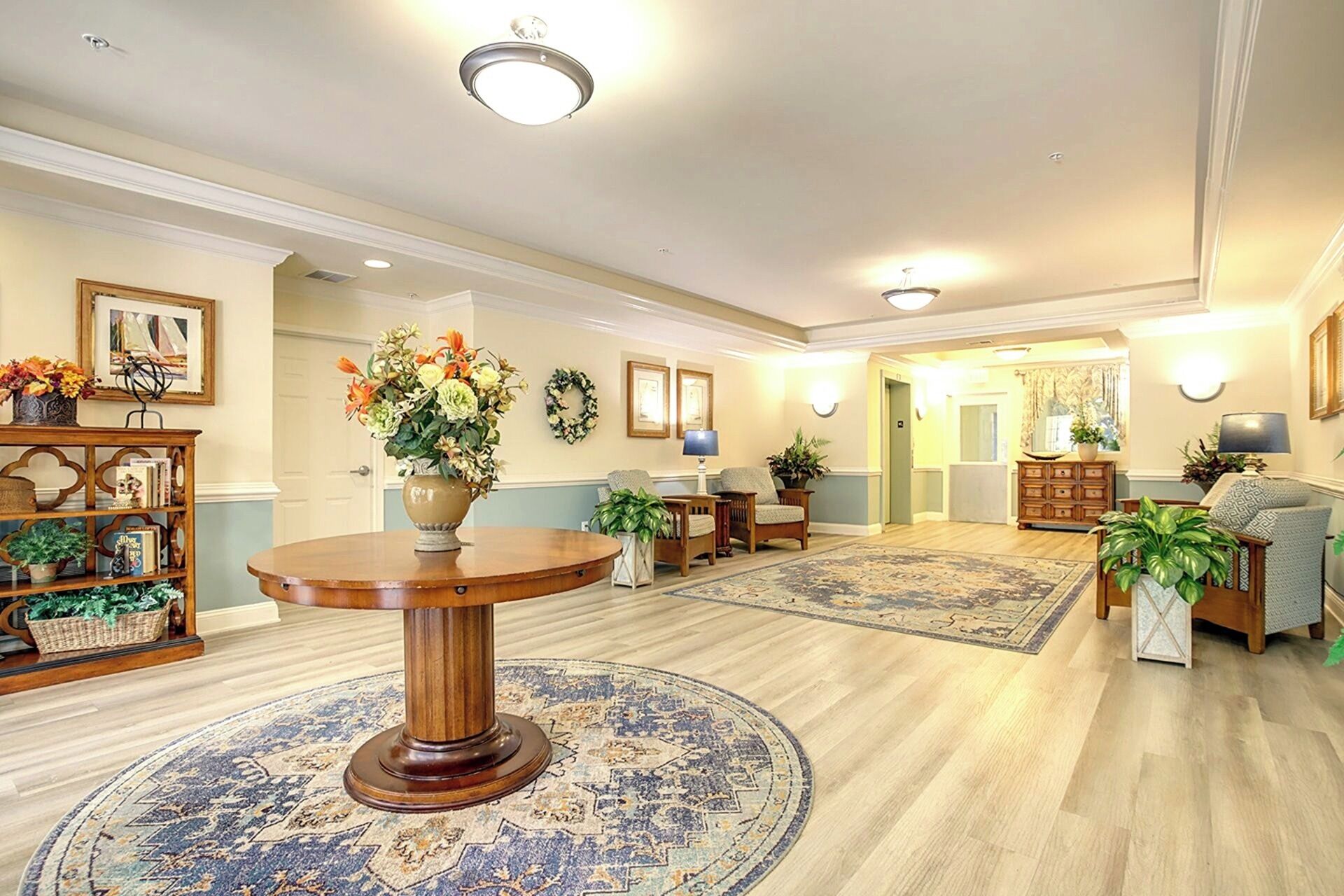 A spacious lobby with a round wooden table, two armchairs, a bookshelf, and a patterned rug.