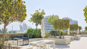 Ein Terrassenbereich im Freien mit Tischen und Stühlen, umgeben von Grünpflanzen und modernen Gebäuden im Hintergrund.