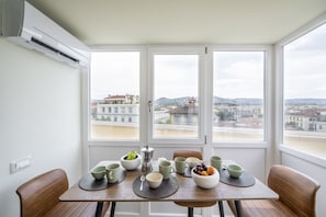Uma mesa de jantar com tigelas, xícaras e uma cafeteira em um cômodo com janelas amplas e um aparelho de ar condicionado.