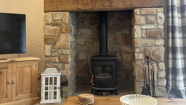 A stone fireplace with a black wood-burning stove, a wooden table with a white bowl, and a white lantern. - Riverside Cottage (Newton Aycliffe)