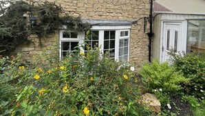A stone house with a white door and windows, surrounded by a garden with various plants and flowers. - Riverside Cottage (Newton Aycliffe)