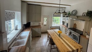 A kitchen with a wooden table, a sink, cabinets, a stove, and a window with a view of greenery. - Riverside Cottage (Newton Aycliffe)