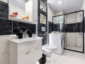 A modern bathroom with a marble backsplash, a sink, a toilet, a washing machine, and a glass-enclosed shower.