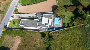 An aerial view of a modern house with a swimming pool, surrounded by lush greenery and a large field.