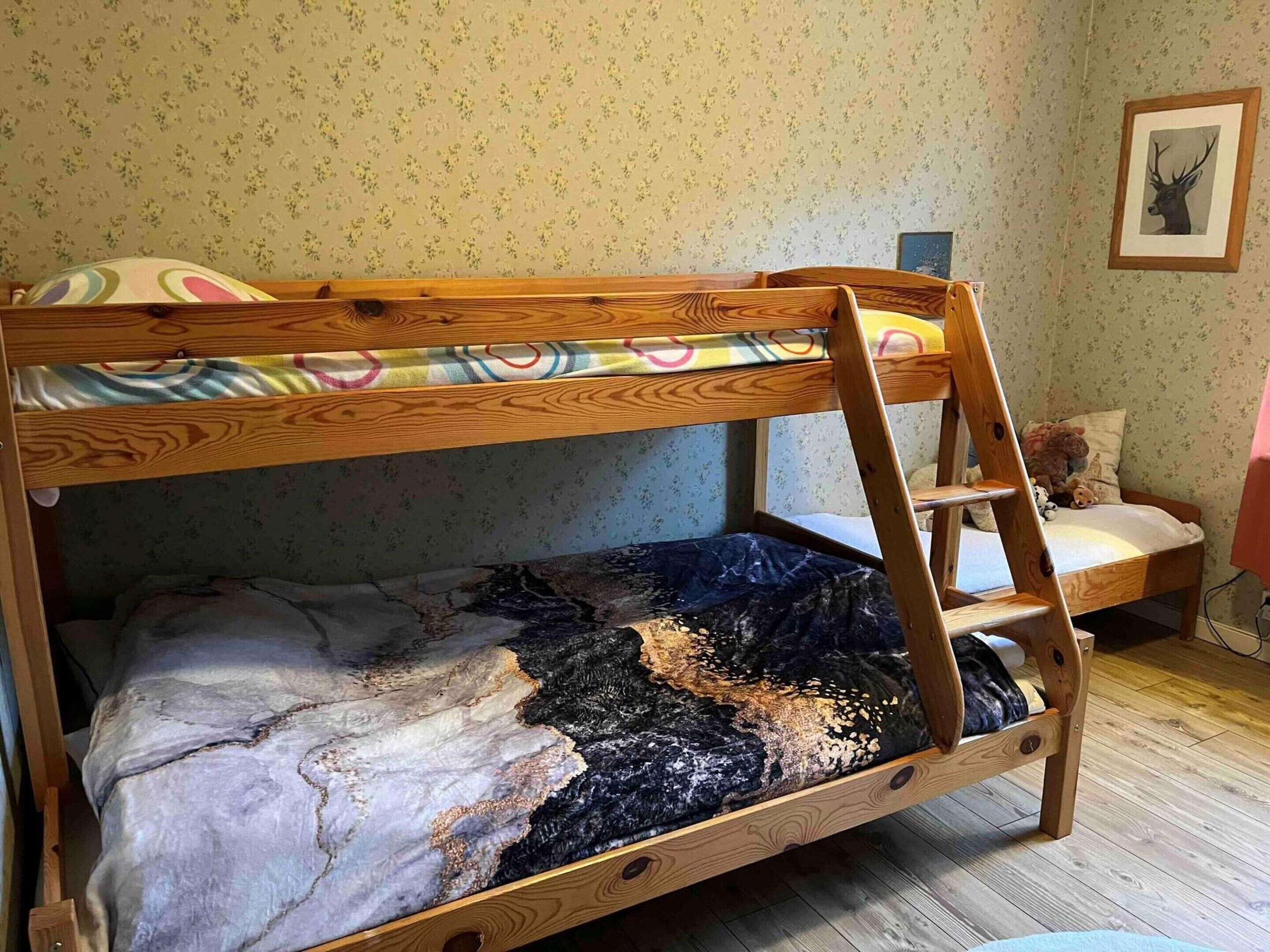 A bunk bed with a wooden frame and a colorful blanket. There is a ladder attached to the side of the bed. The room has floral wallpaper and a framed picture on the wall.