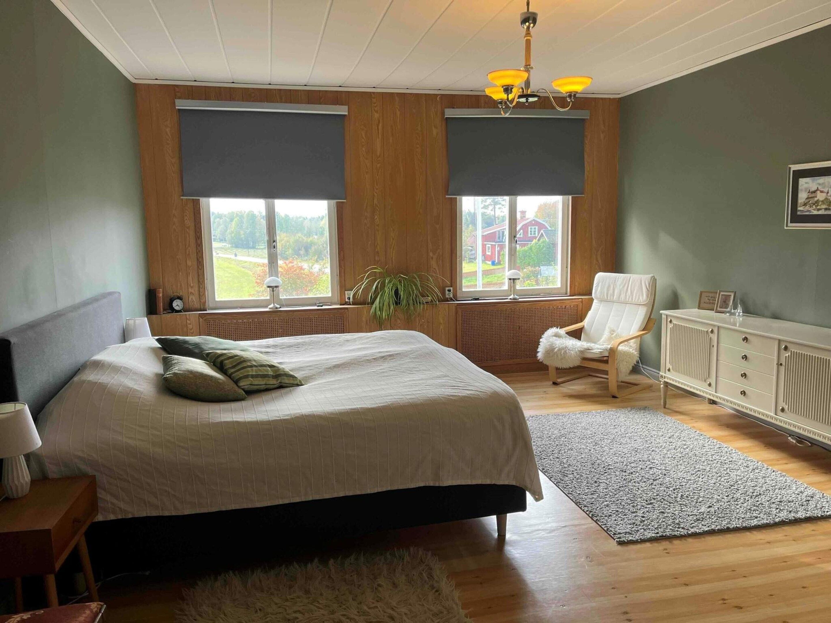 Un dormitorio con cama, una silla, un radiador y una ventana con vista a una casa.
