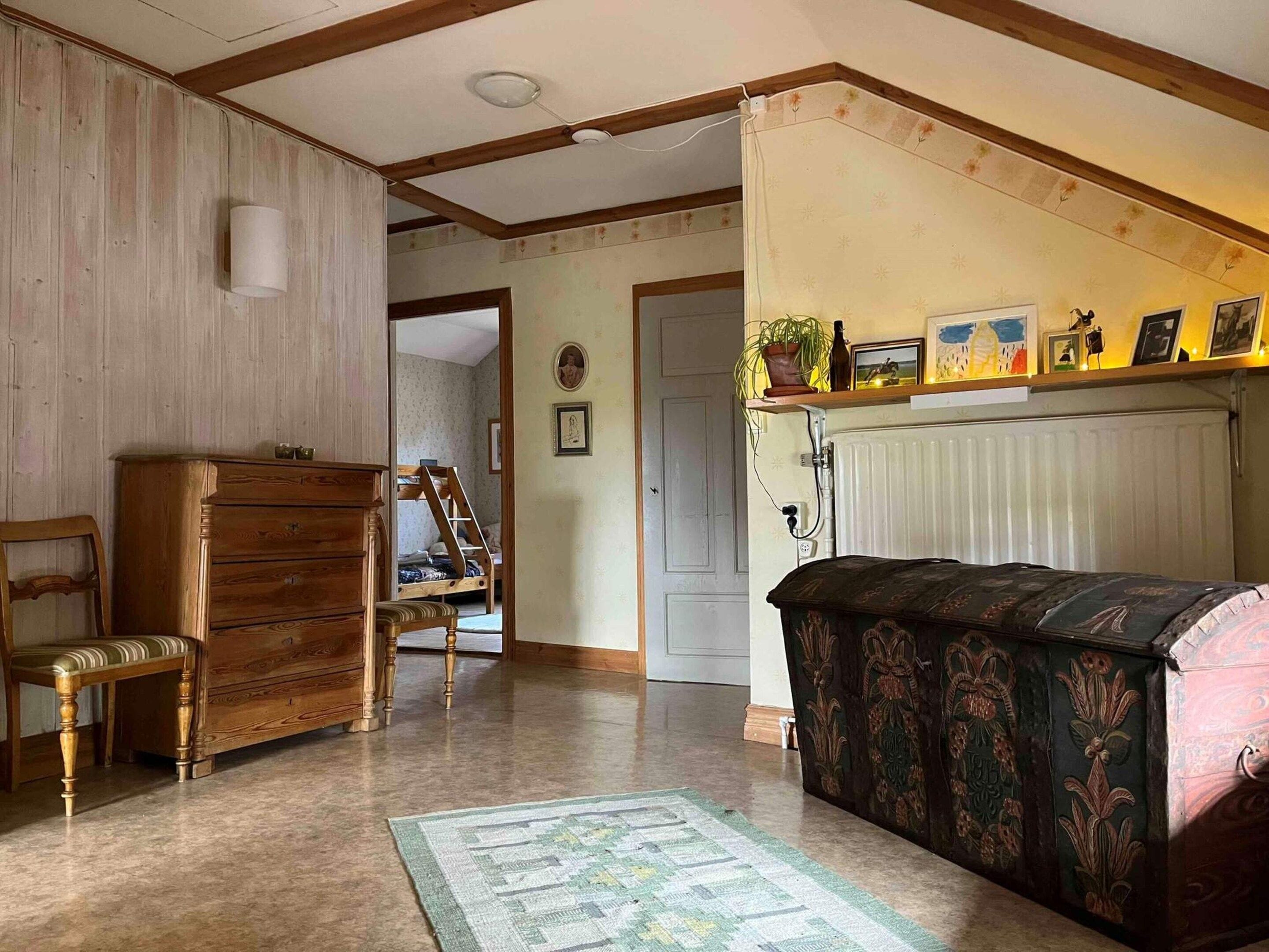 A room with a wooden dresser, a chair, a bed with a patterned cover, a radiator, and a shelf with pictures and a plant.