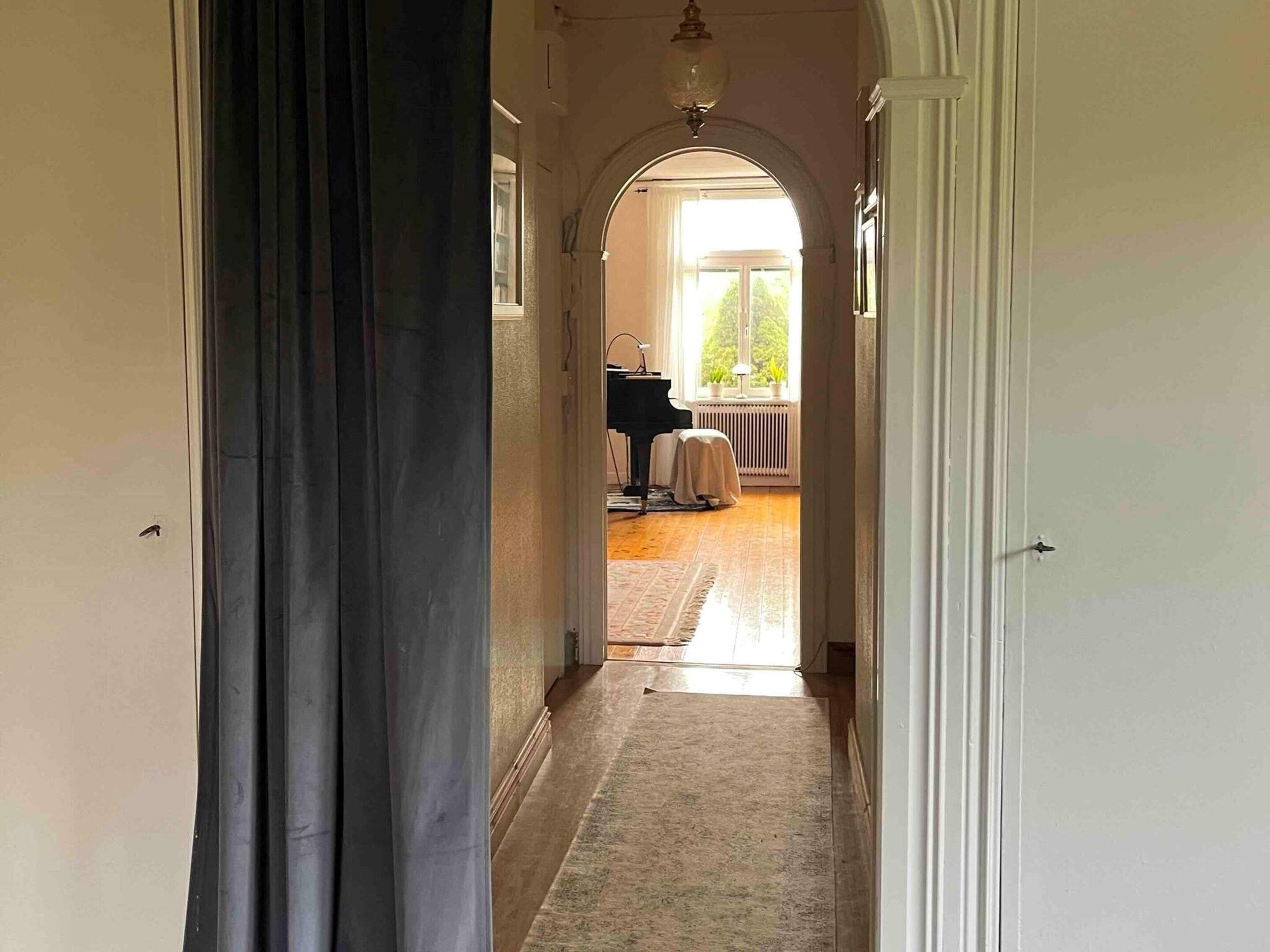 A hallway leading to a room with a piano, a radiator, and a view of the outdoors.
