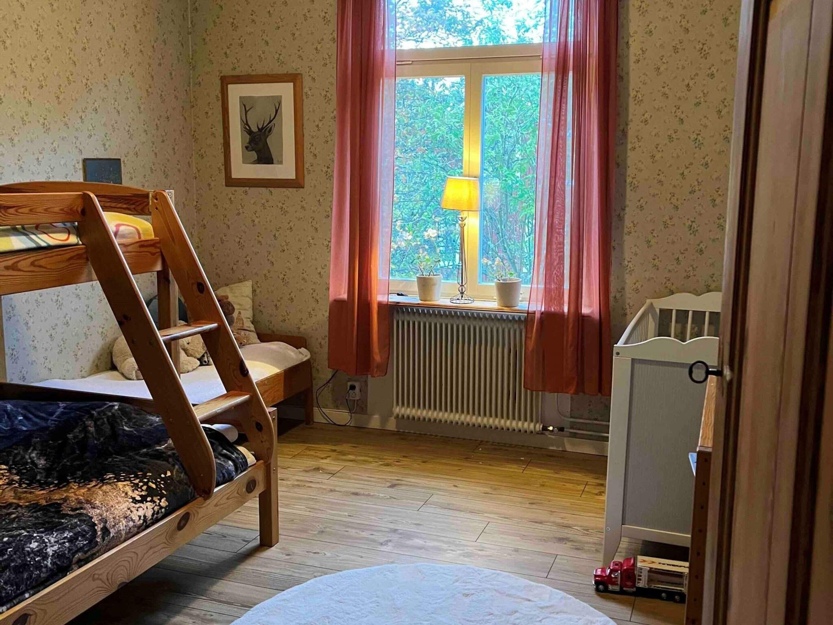 A child's bunk bed with a wooden ladder, a small bedside table, a lamp, and a window with curtains.
