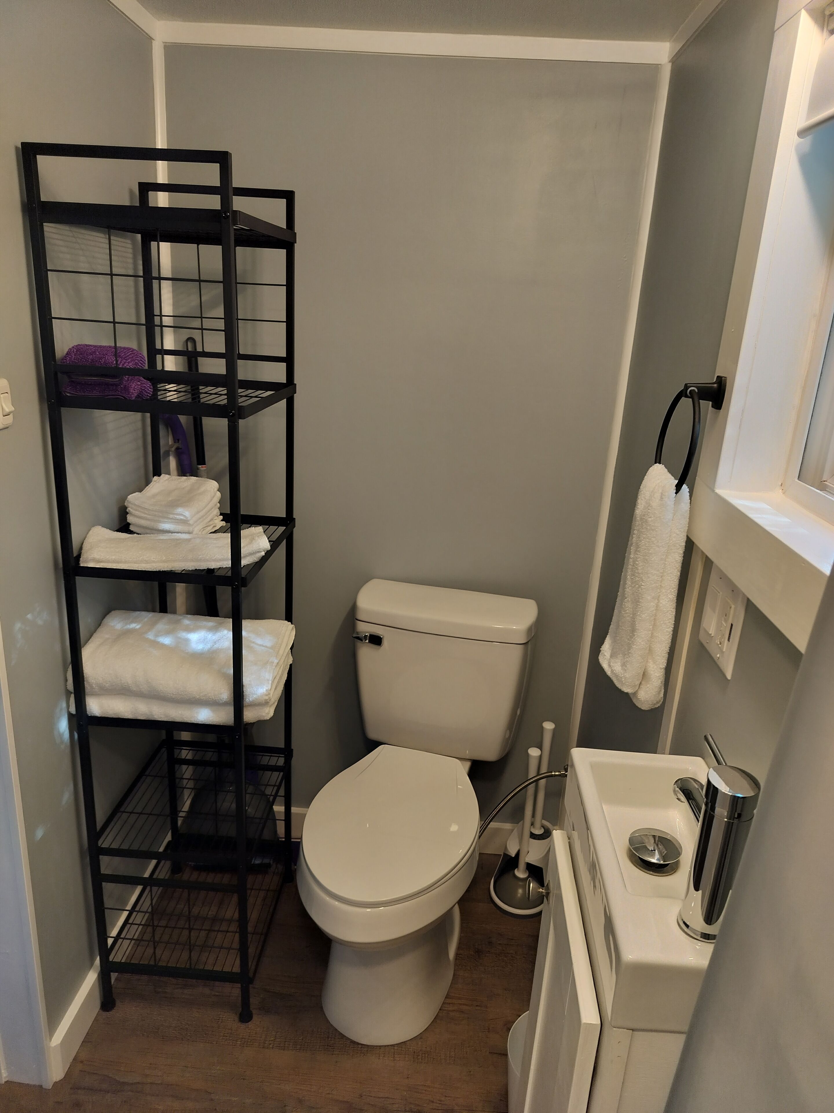 Une petite salle de bain avec des toilettes blanches, une étagère en métal noir où sont posées des serviettes, et un lavabo blanc avec un miroir.