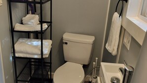 Une petite salle de bain avec des toilettes blanches, une étagère en métal noir où sont posées des serviettes, et un lavabo blanc avec un miroir.
