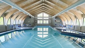 Una piscina interior con agua azul cristalina, vigas de madera en el techo y zonas de estar con mesas y sillas.