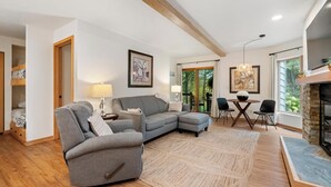 Una sala moderna con un sofá seccional gris, piso de madera y chimenea. Hay un comedor con mesa y sillas, y un ventanal que ofrece vistas a la vegetación exterior.