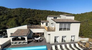 A modern multi-story building with a pool, outdoor seating, and a view of a forested hillside.