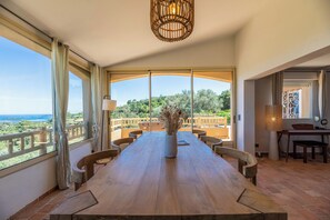 A spacious dining area with a long wooden table, multiple chairs, and a vase of dried flowers. Large windows offer a view of the outdoors. - Luxury villa with exceptional panoramic views in Porto Vecchio (Porto-Vecchio)