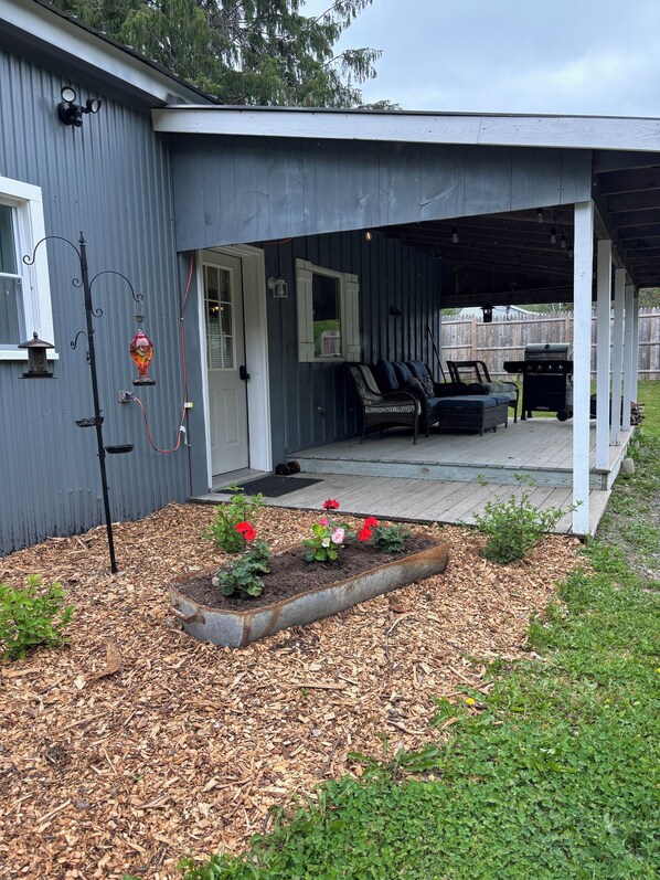 A covered porch with outdoor seating, a garden bed with flowers, and a grill.