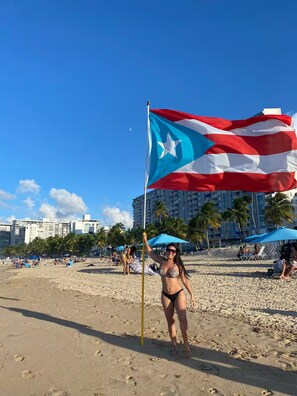 A beach scene with a flag, palm trees, and buildings in the background. - Casa Dos Palmas-House with Solar System and just 8 Min from the Airport. (San Juan)