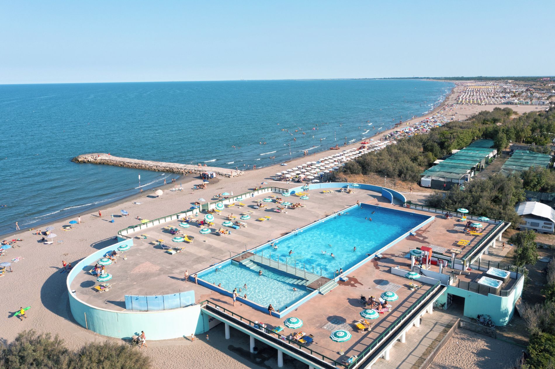Une grande piscine entourée de chaises longues et de parasols, avec une plage et l’océan en arrière-plan.