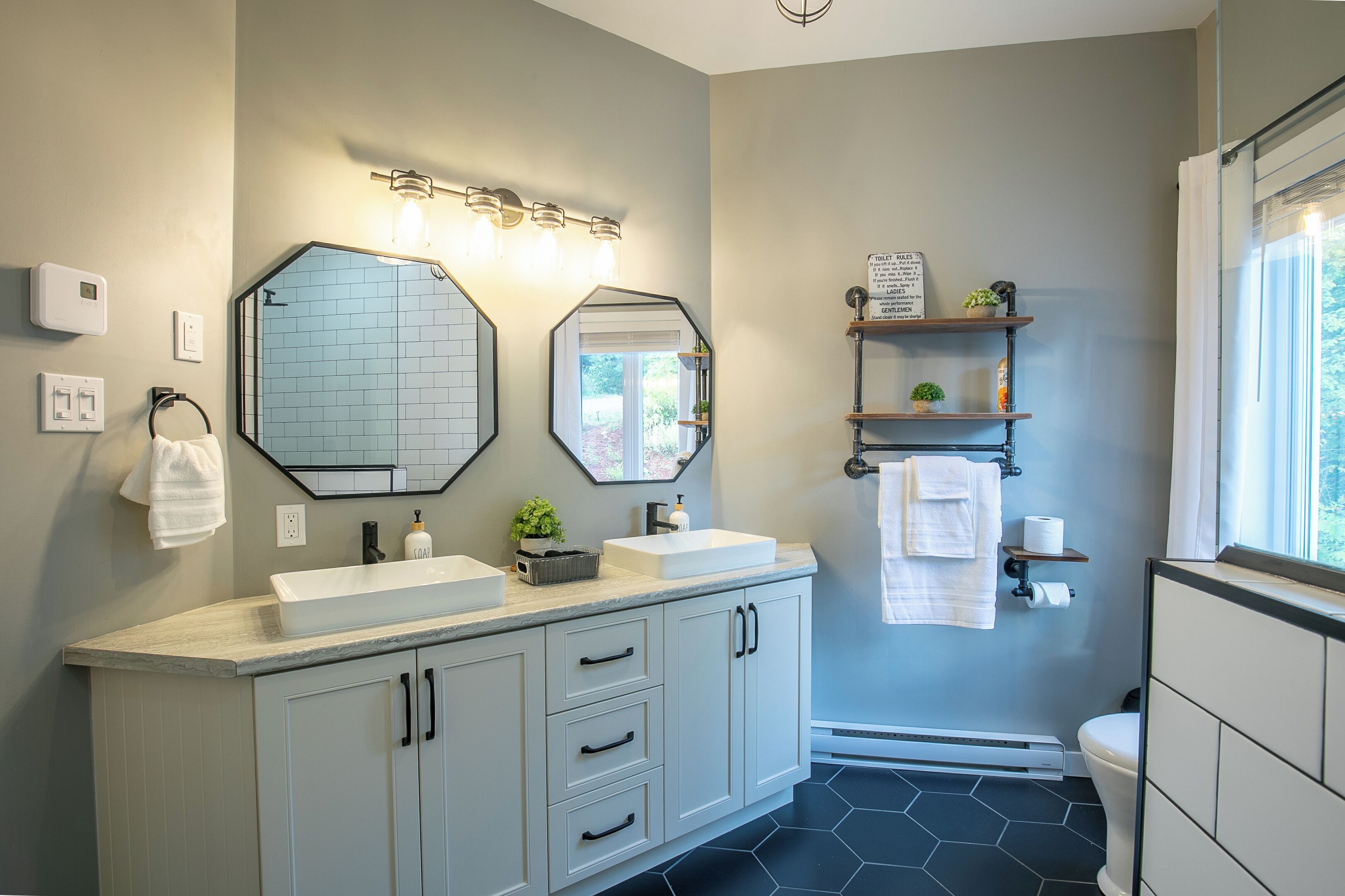 A bathroom with two sinks, a toilet, and a shower. There are mirrors, a towel rack, and a shelf with toiletries.
