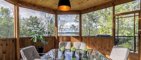 Puupaneloitu huone, jossa on lasipöytä ja -tuolit, riippuvalaisin sekä suuret ikkunat, joista avautuu näkymä puihin ja järvelle.