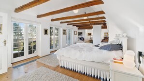 Une chambre avec un grand lit, des poutres en bois, une commode blanche et une vue sur l’extérieur.