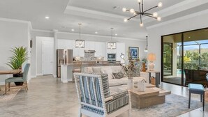 A spacious living area with a modern sofa, a patterned armchair, and a coffee table. A kitchen with white cabinets and a dining area with a table and chairs are visible in the background. - Looking for last minute X-mas plans?  Beautiful home in Fort Myers - 7 days min. (Fort Myers)
