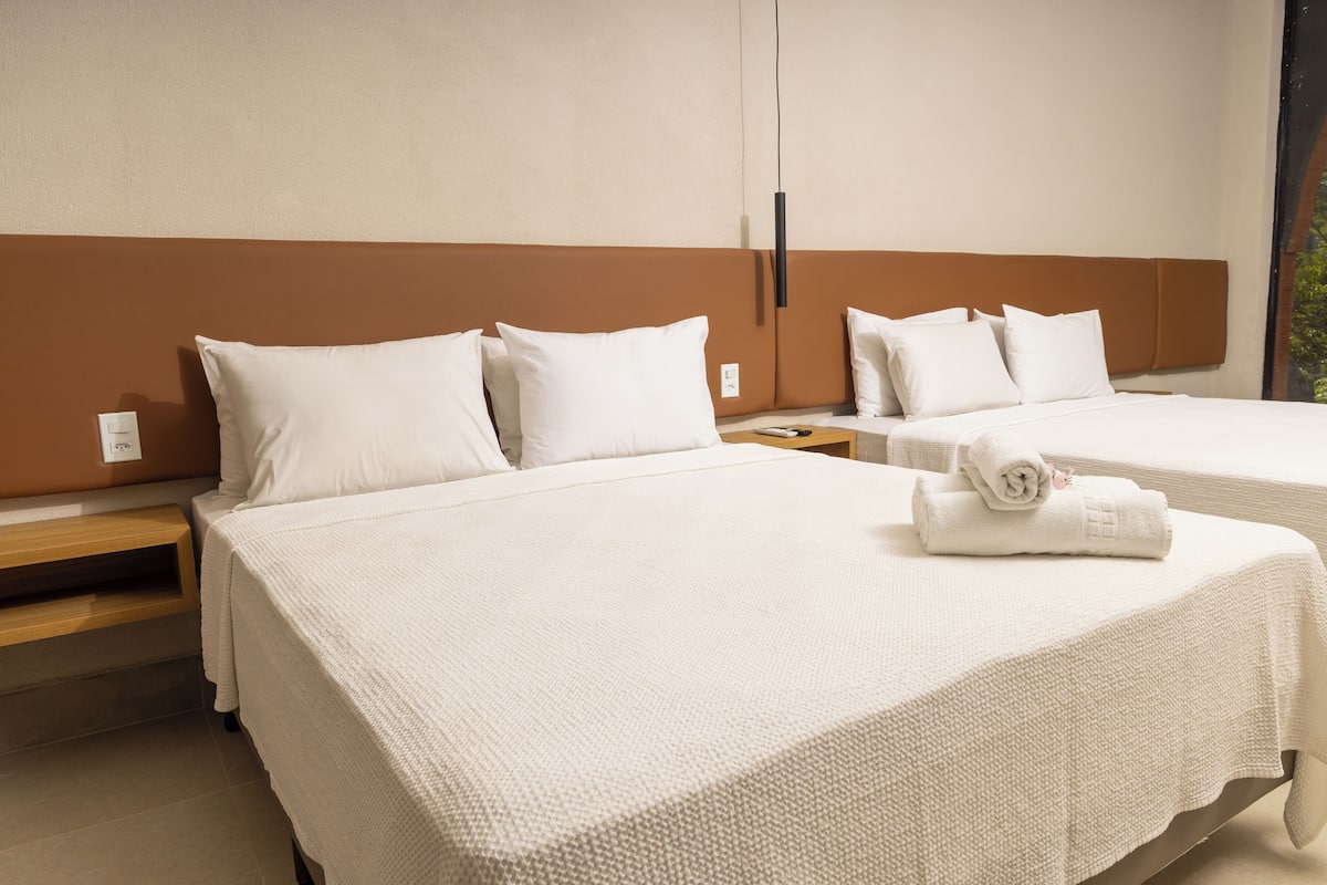 A hotel room with two beds, each with white bedding and pillows, a wooden nightstand, and a view of greenery outside.