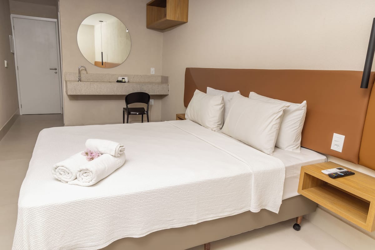 A hotel room with a large bed, a bedside table, a mirror, a small wooden shelf, and a bathroom area with a sink and chair.