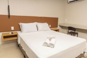 Una cama bien hecha con sábanas blancas, un par de toallas enrolladas, una mesita de noche con un control remoto y una silla en una habitación de hotel.