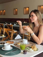Una persona sentada a una mesa con un plato que incluye tostadas, una bebida verde y un postre pequeño, con un sujetapapeles y una taza de té a la vista.