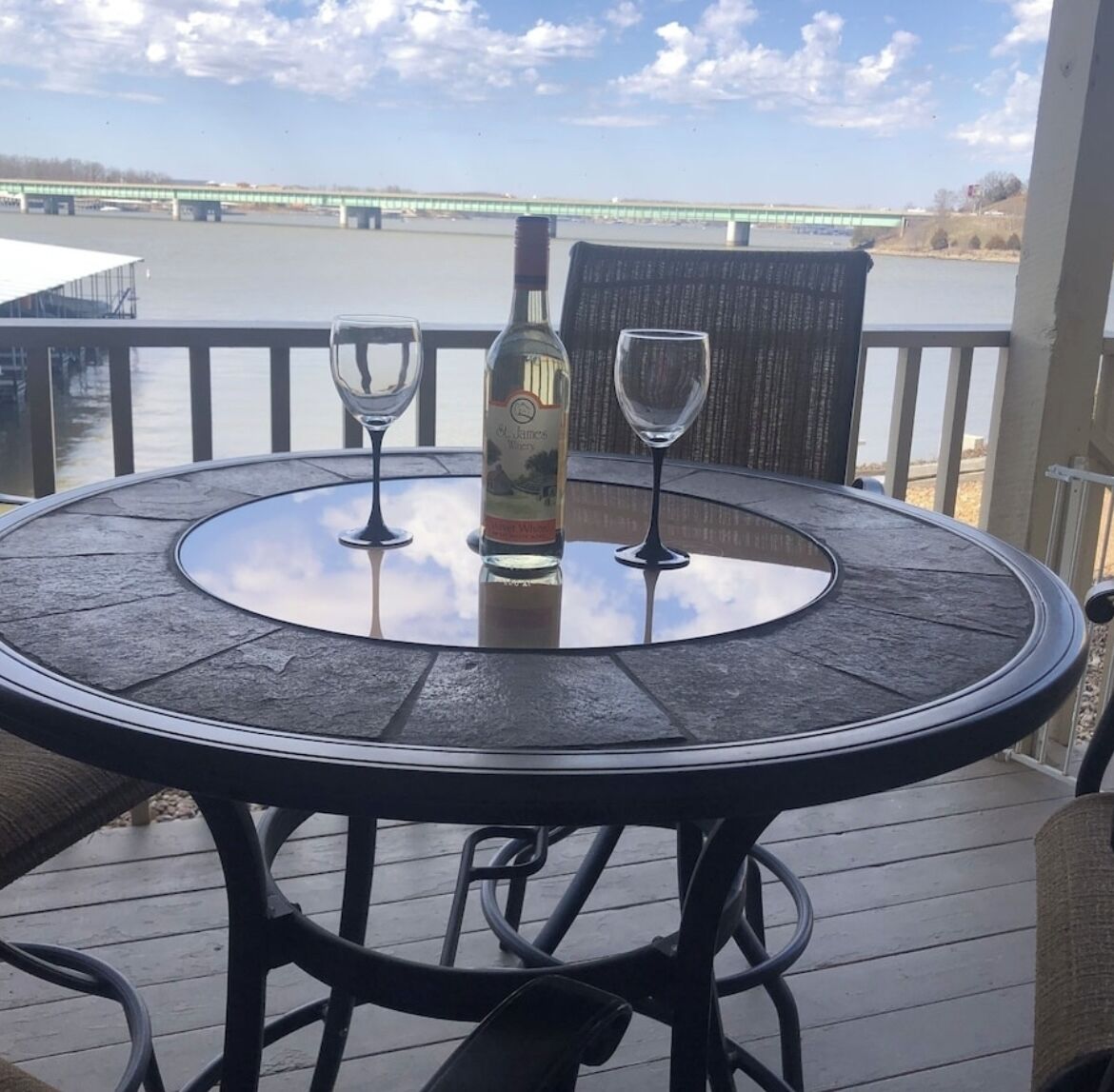 A round table with a bottle and two glasses on a deck with a view of a bridge.