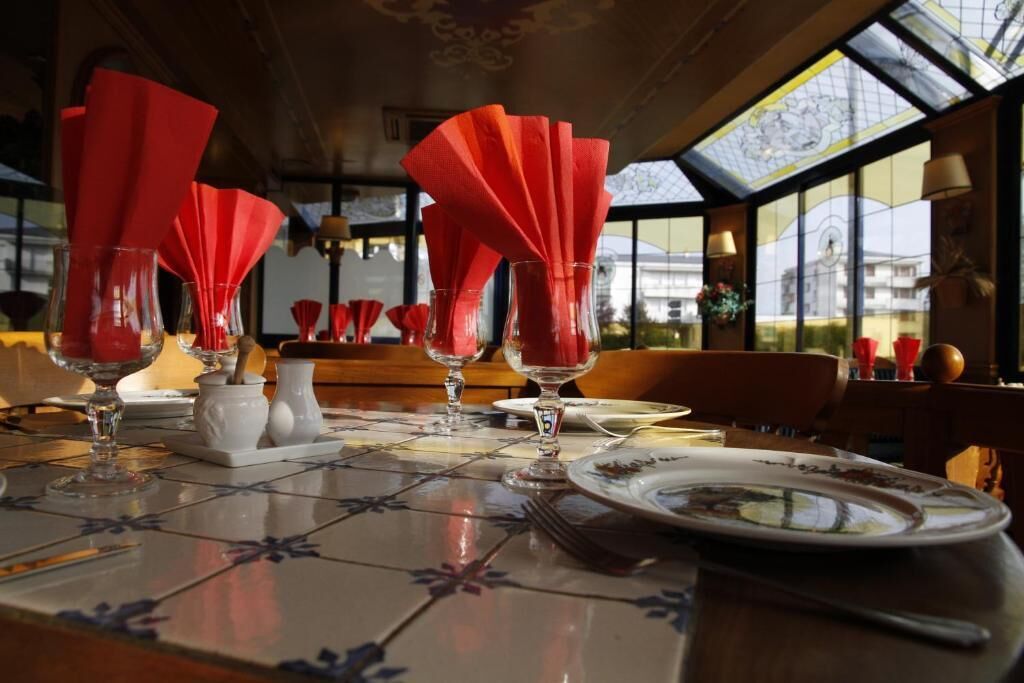 Mesa de comedor servida con servilletas rojas, copas de vino y platos, con vista al exterior a través de amplios ventanales.