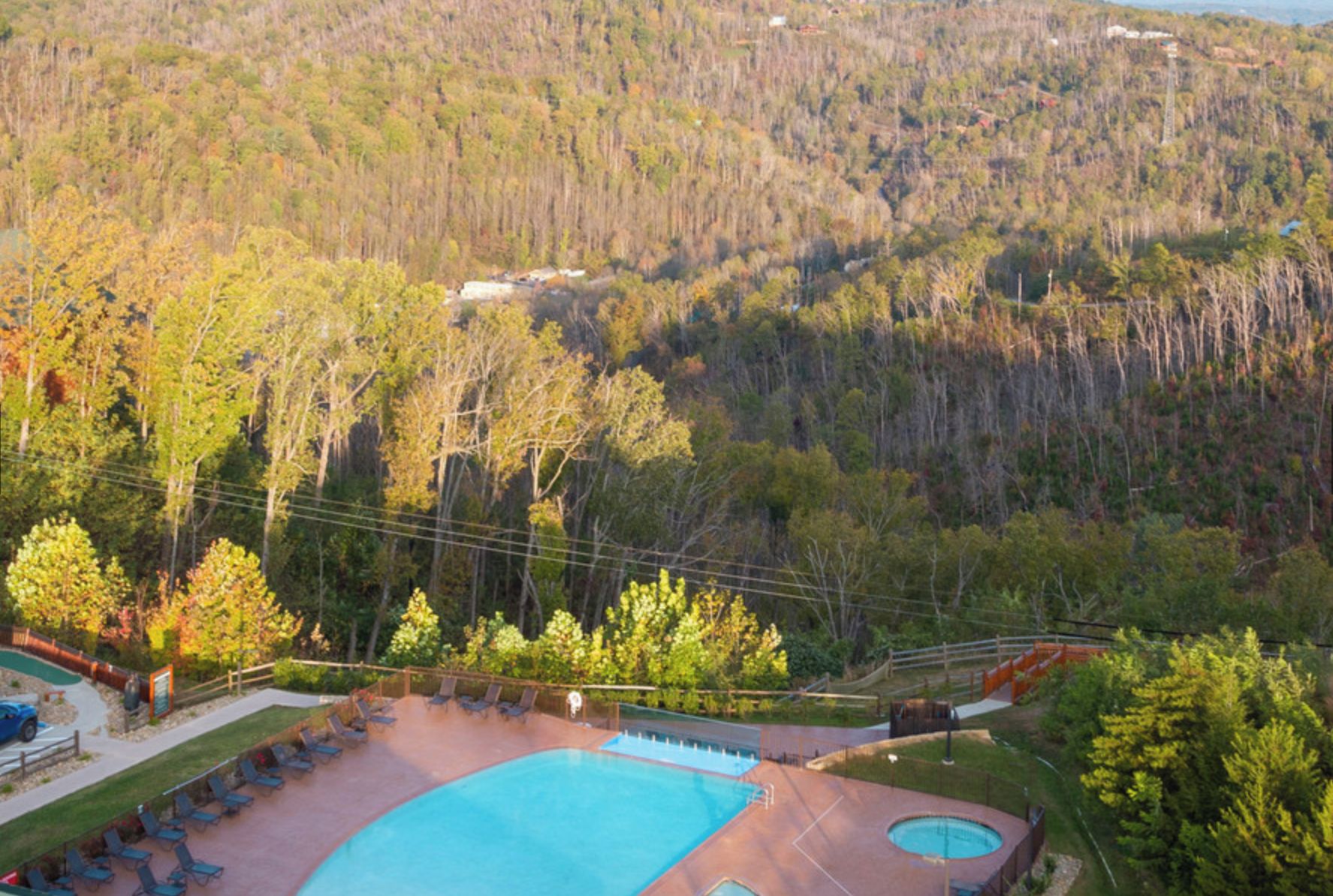 Vista aérea de un resort con piscina, sillas de descanso y un paisaje boscoso de fondo.