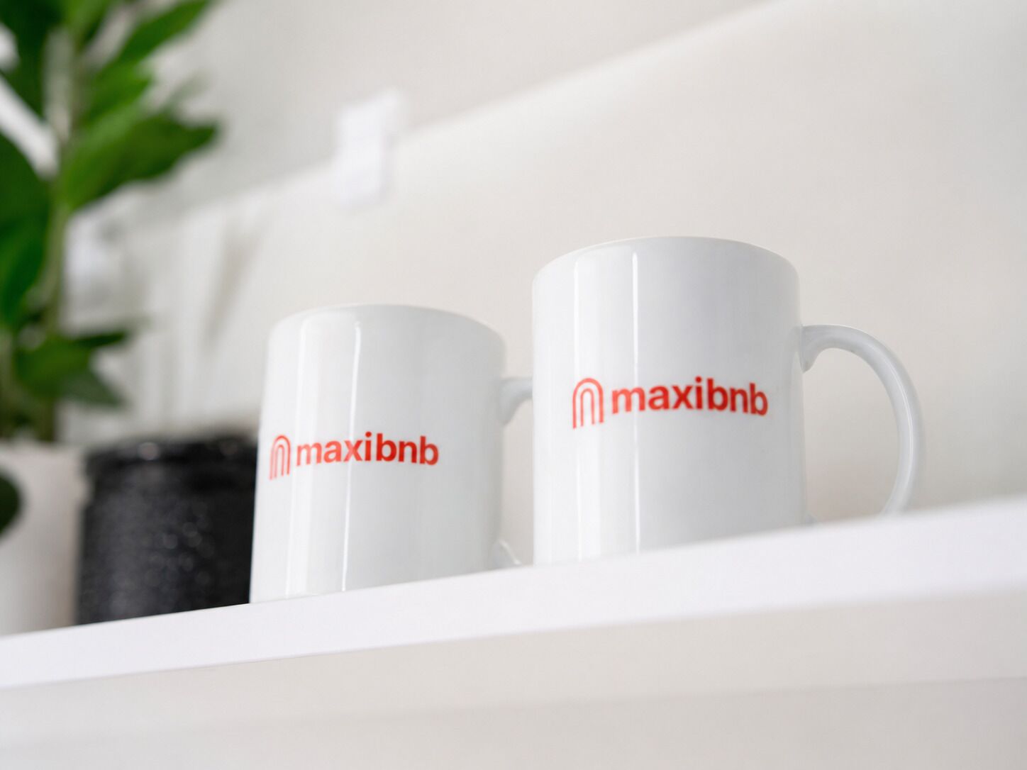 Dos tazas blancas con el logotipo 'maxibnb' sobre un estante blanco.