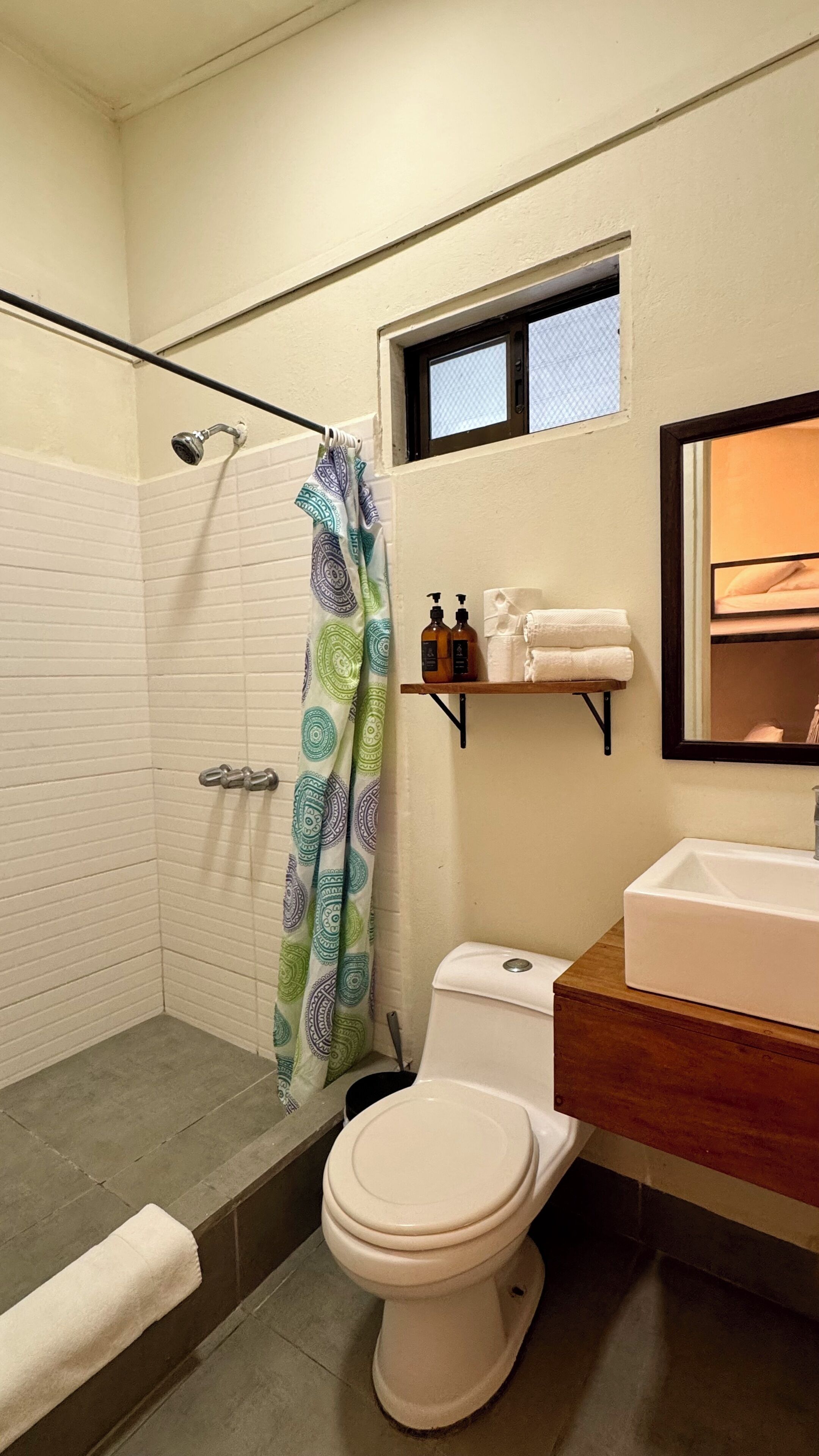 A bathroom with a shower, toilet, sink, and a small window above the mirror.