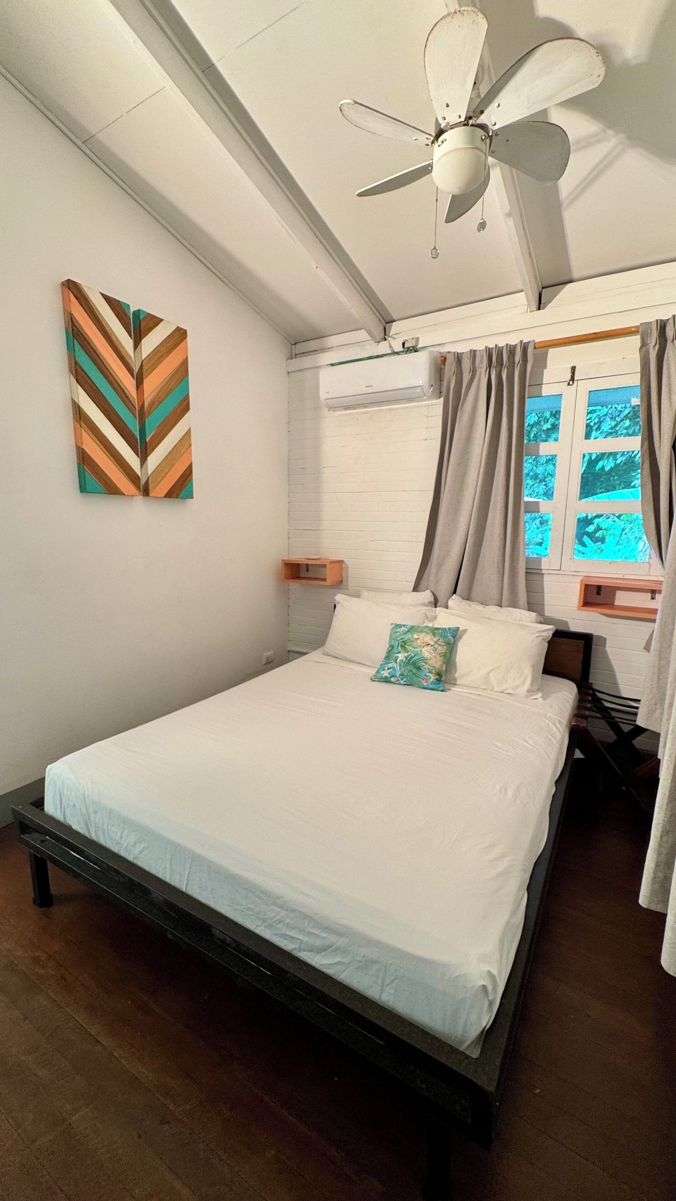 A neatly made bed with white linens, a ceiling fan, a window with curtains, and a wall-mounted air conditioner.