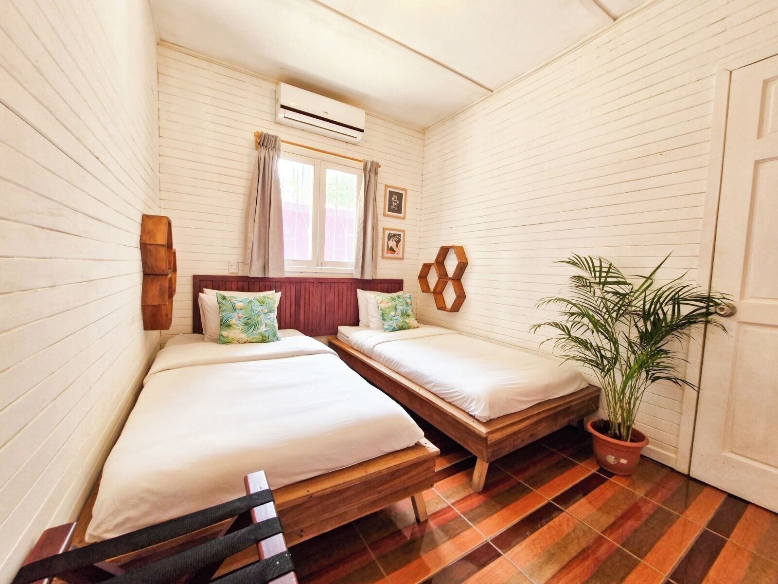 A small, cozy room with two beds, a wooden bench, a potted plant, and a window with curtains.