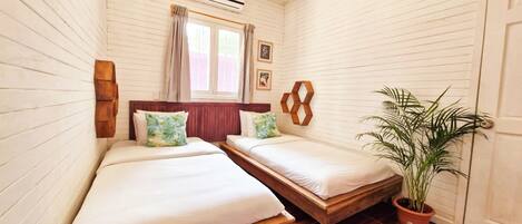 A small, cozy room with two beds, a wooden bench, a potted plant, and a window with curtains.