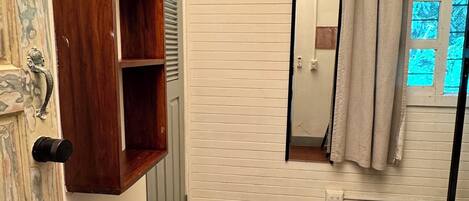 Une pièce avec un banc en bois, un climatiseur blanc, une porte avec un miroir et une fenêtre avec un rideau.