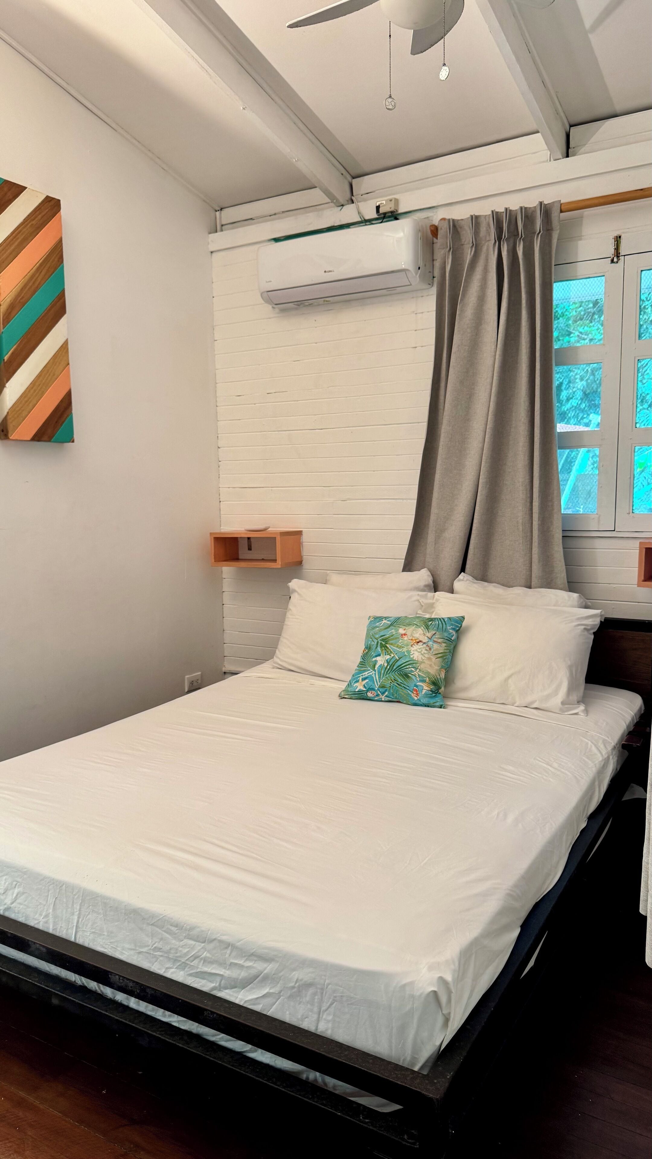 Een slaapkamer met een bed, witte muren, een plafondventilator, een airconditioning en een raam met gordijnen, plus een houten plank.