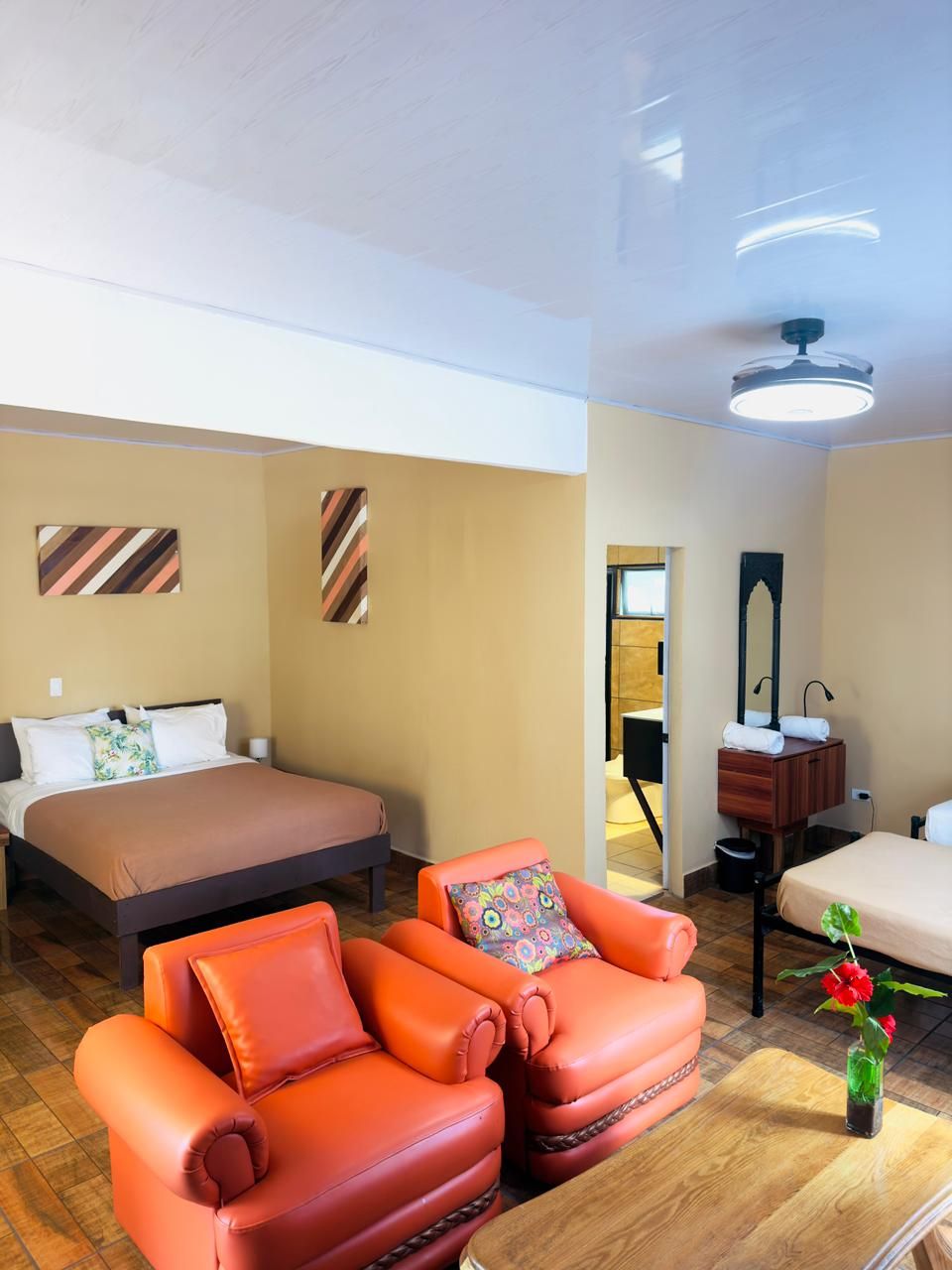 Habitación de hotel con una cama, dos sillones naranjas, una mesa de madera, un espejo y una lámpara en el techo.
