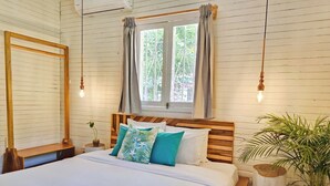 A bedroom with a wooden bed frame, white bedding, and a window with curtains. There are two bedside tables with lamps, a potted plant, and a wooden stool. - Agapi (Cahuita)