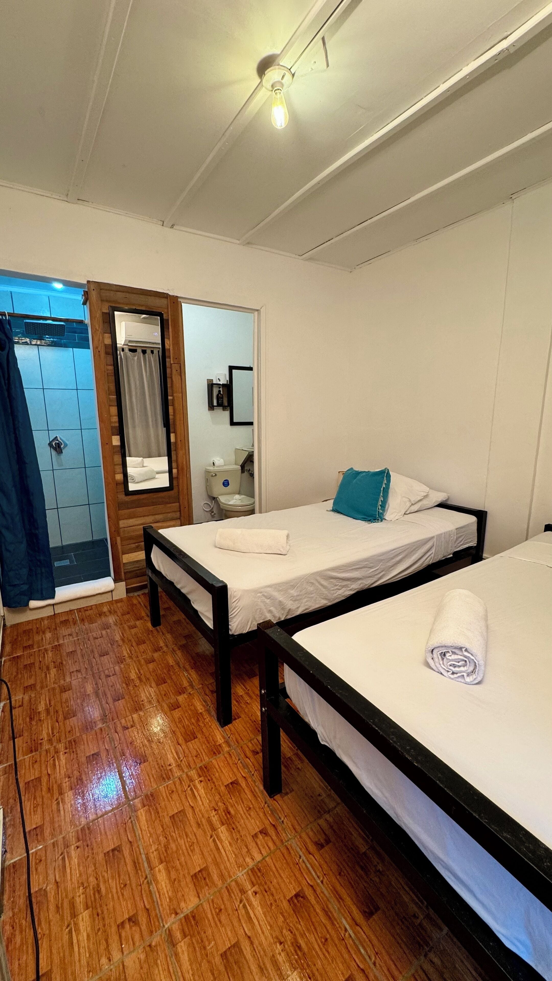 A room with two beds, a wooden floor, a ceiling fan, and a bathroom visible through an open door.