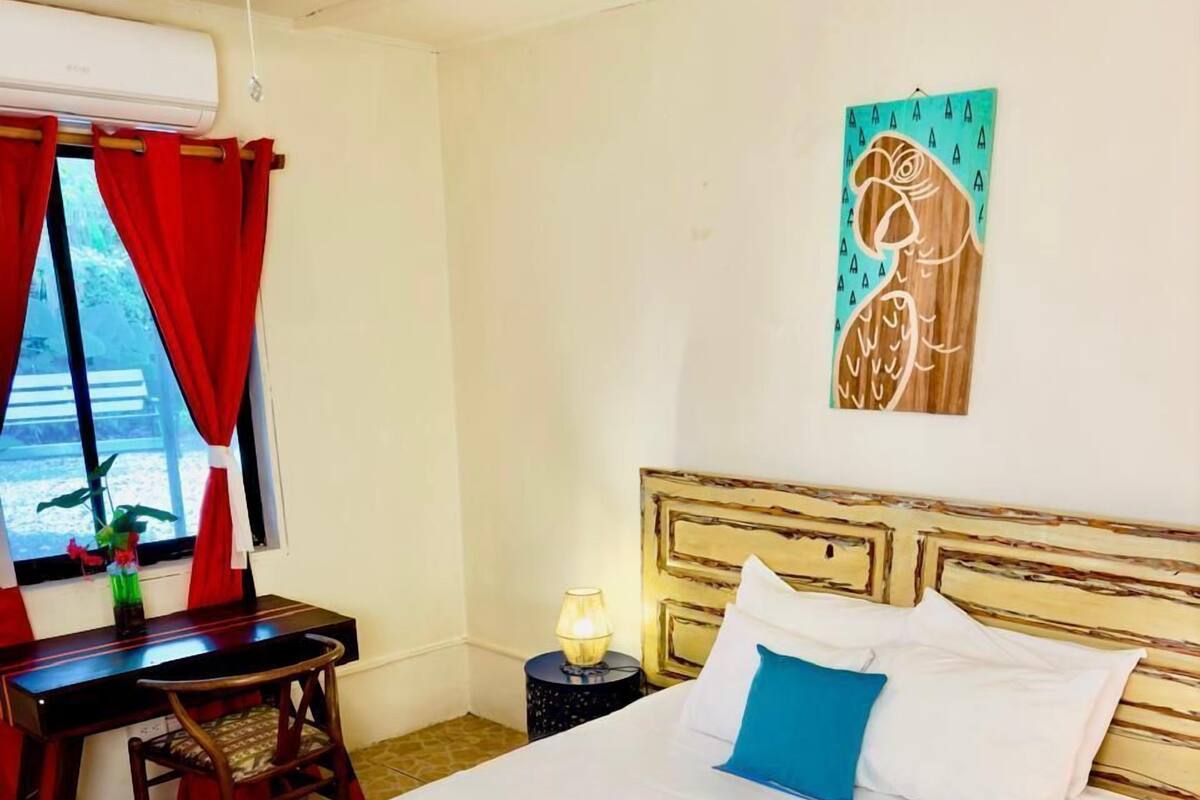 Habitació d'hotel amb un llit gran, escriptori, cadira, tauleta de nit amb una làmpada i finestra amb cortines vermelles.