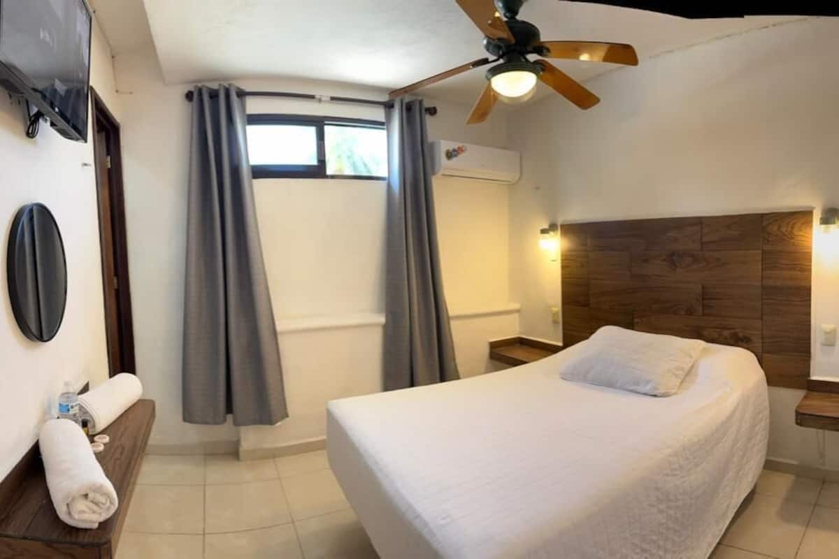 A hotel room with a bed, a ceiling fan, a TV, a wooden headboard, and a window with curtains.